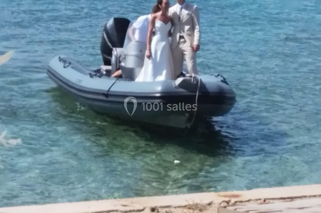 Un couple en tenue de mariage se tient debout sur un bateau pneumatique amarré près d'une plage.