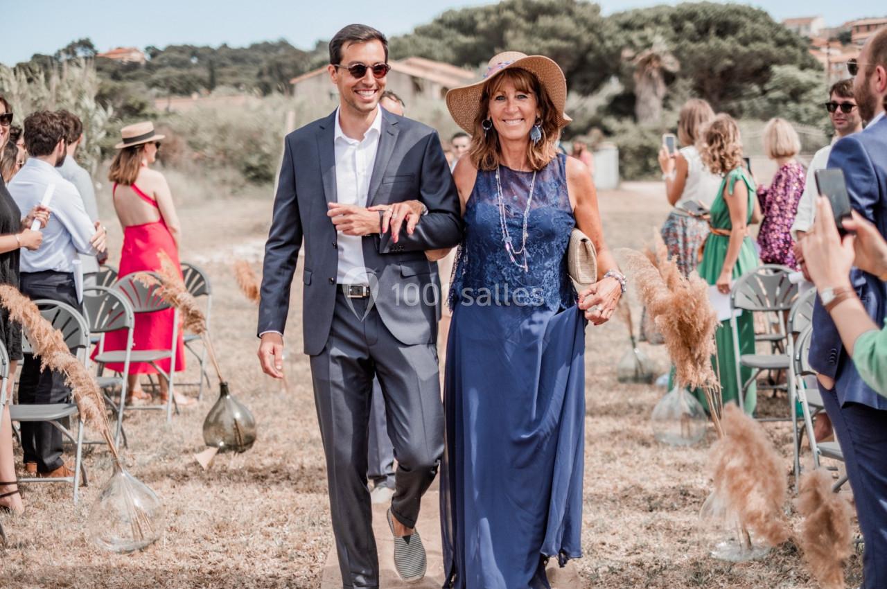 Un homme en costume et une femme en robe bleue avancent ensemble lors d'une cérémonie en extérieur.
