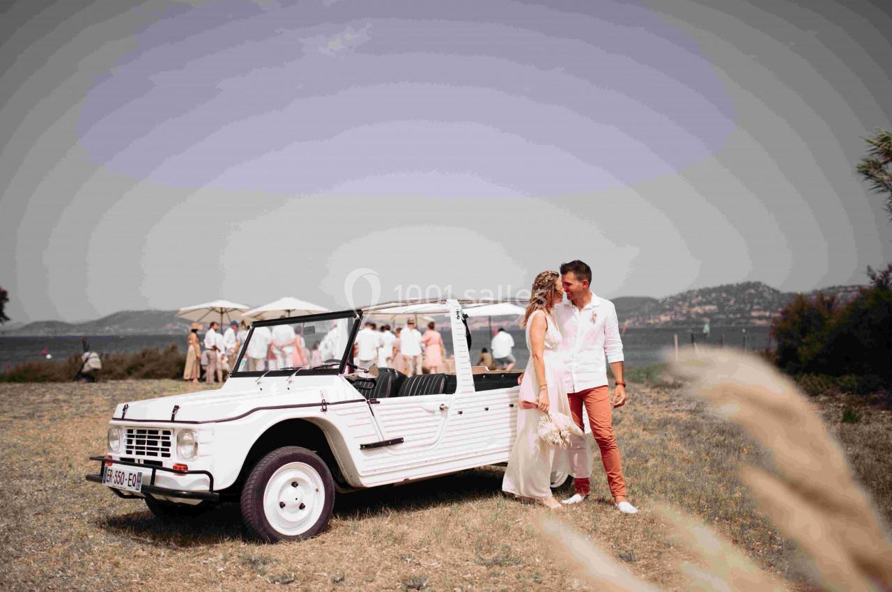 Un couple debout près d'une voiture décapotable blanche dans un champ, avec un groupe de personnes en arrière-plan.