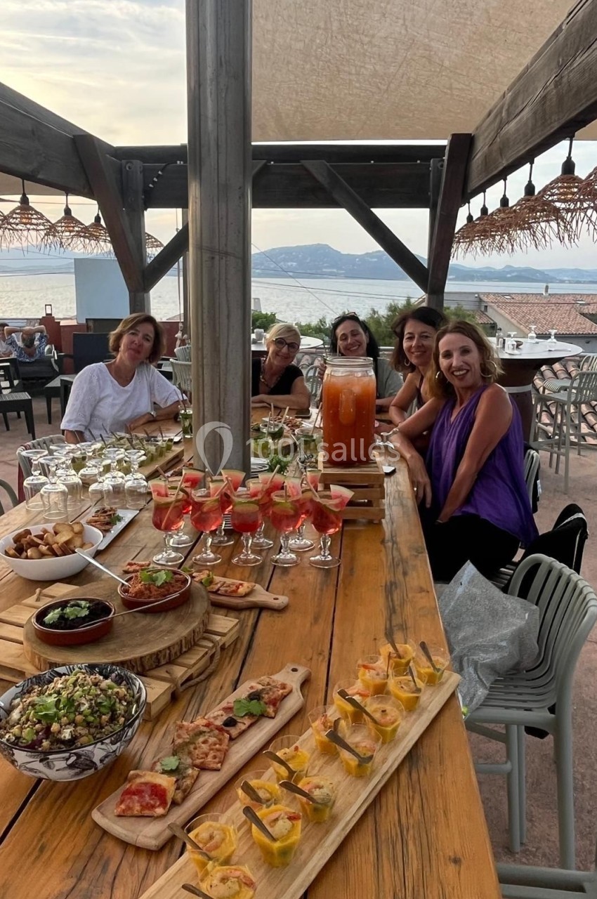Un groupe de personnes assises autour d'une table en bois garnie de plats et boissons, en terrasse avec vue sur la mer.