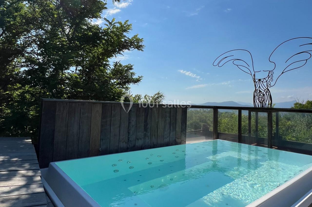 Bain à remous extérieur sur une terrasse en bois, entouré de verdure, avec deux verres de vin et vue dégagée sous un ciel…