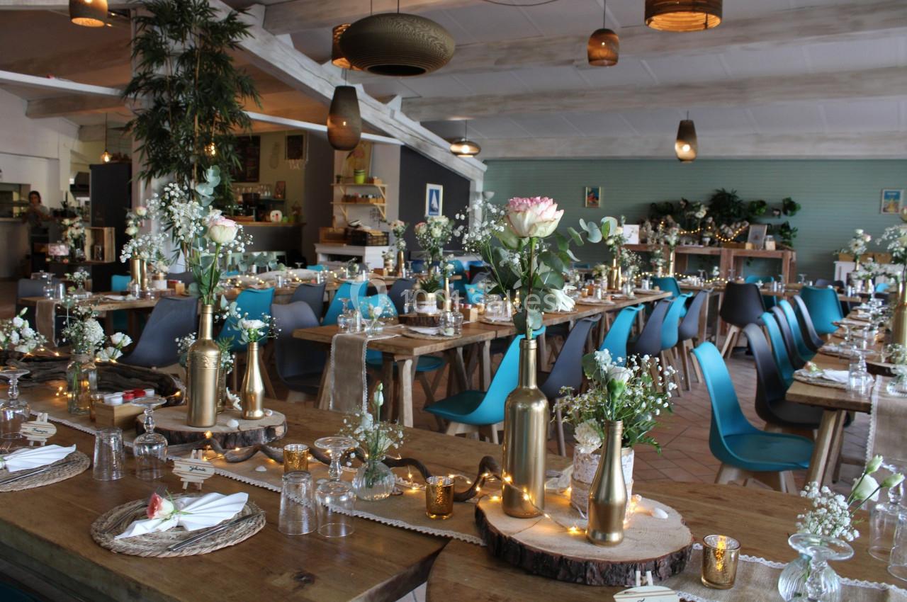 Salle de réception décorée pour un événement, avec tables en bois ornées de fleurs, bougies et éléments dorés.