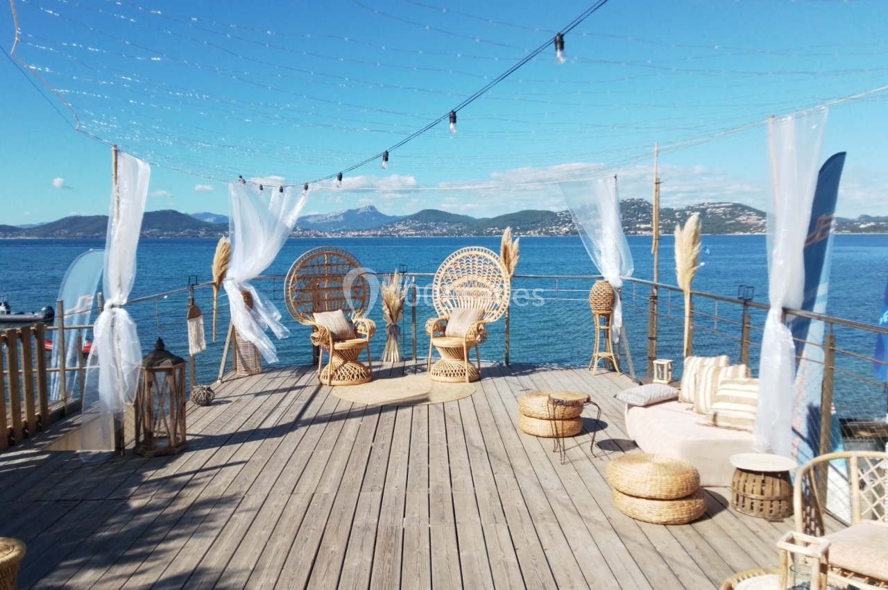 Terrasse en bois avec mobilier en rotin, vue sur la mer, montagnes en arrière-plan et ciel dégagé.
