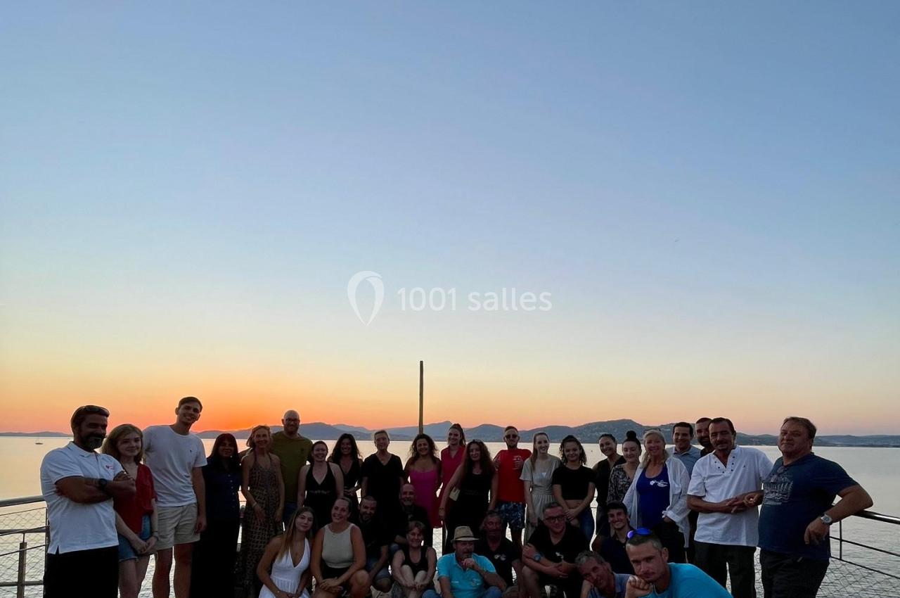 Groupe de personnes posant sur une terrasse en bois au coucher du soleil avec une vue sur la mer et des montagnes.