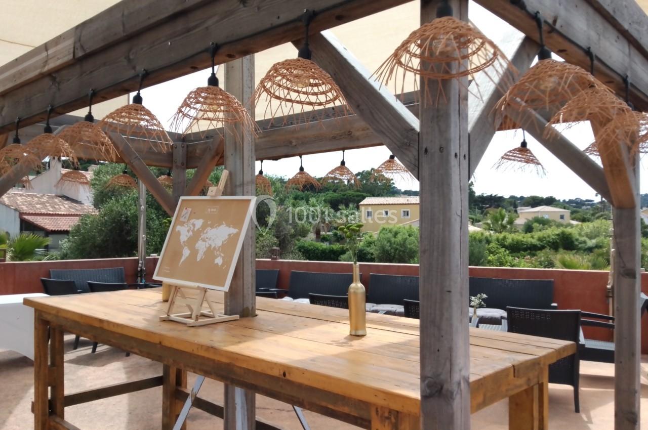 Table en bois sous une pergola avec suspensions en osier, carte du monde posée dessus, vue sur des jardins en arrière-plan.