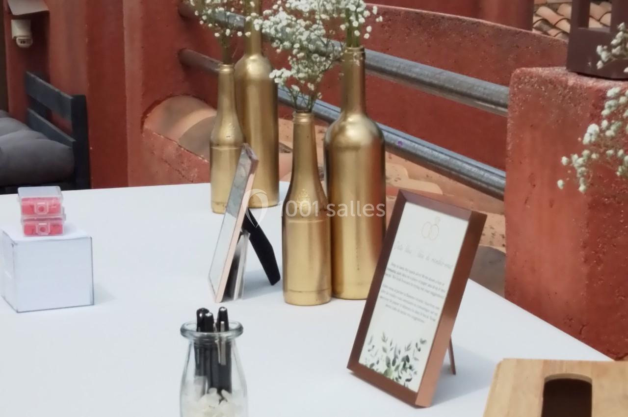 Table décorée avec des vases dorés contenant des fleurs, sur une terrasse avec vue sur la mer et des toits en arrière-plan.