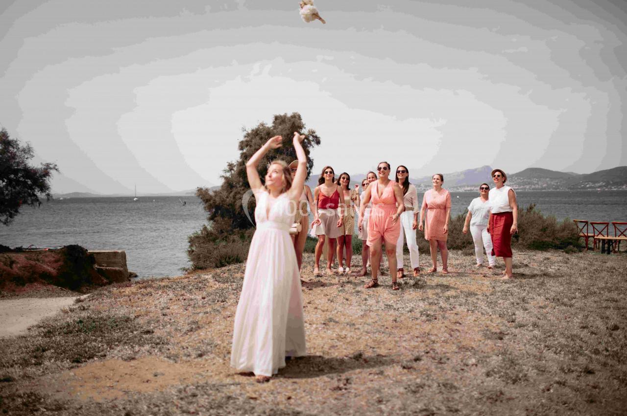 Une mariée lance son bouquet en direction d'un groupe de femmes sur un terrain herbeux près d'un plan d'eau.