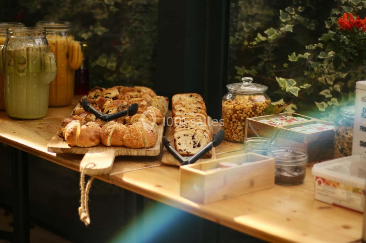 Location salle Hyères (Var) - La Réserve Hyères - Giens #7 Table en bois avec viennoiseries, pain aux raisins, pots de céréales et boissons dans un cadre végétalisé.