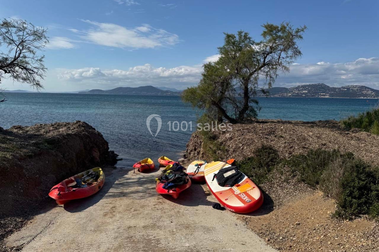 Location salle Hyères (Var) - La Réserve Hyères - Giens #2 Des kayaks et une planche de paddle posés sur une rampe en béton menant à la mer, avec un paysage côtier en arrière-plan.
