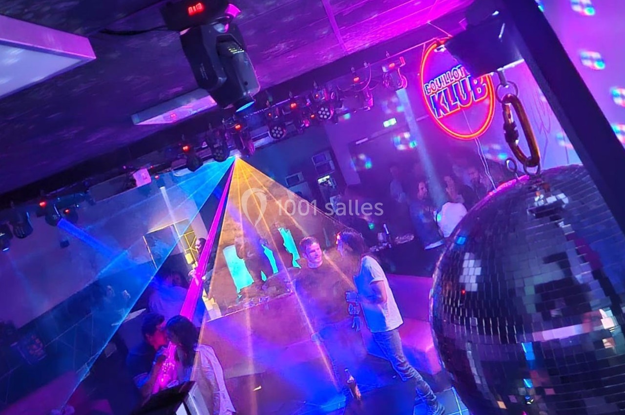 Location salle Hyères (Var) - La Réserve Hyères - Giens #11 Des personnes dansent dans une boîte de nuit avec des jeux de lumière colorés, un miroir disco et un bar en arrière-plan.