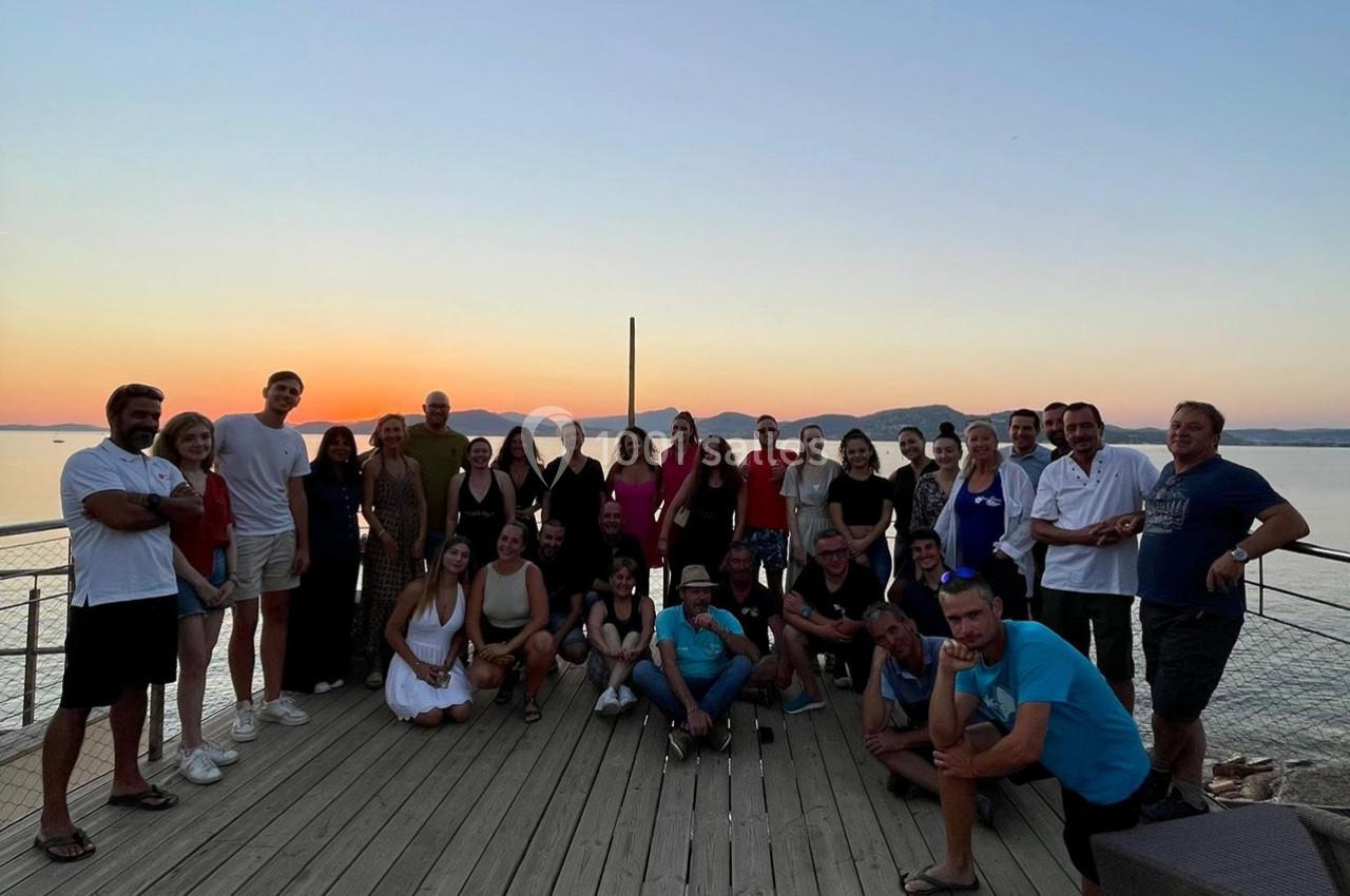 Cohésion sur la Pointe Panoramique - Restaurant Le Solarium Groupe de personnes posant sur une terrasse en bois au bord de l'eau, avec un coucher de soleil en arrière-plan.