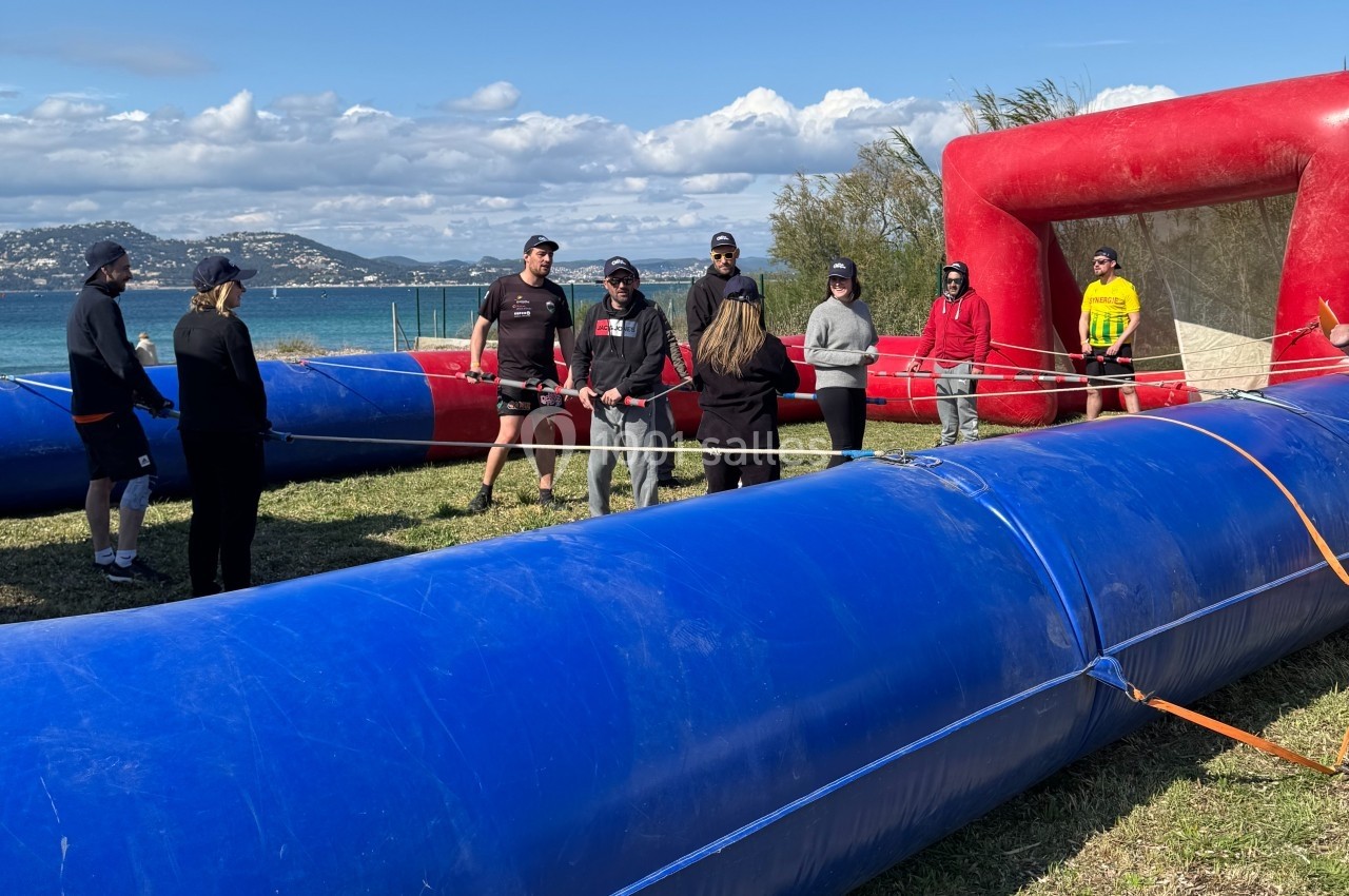 Cohésion sur le Terrain Les Pieds dans l'Eau Des personnes debout autour d'un terrain gonflable bleu et rouge en plein air, près d'un plan d'eau.