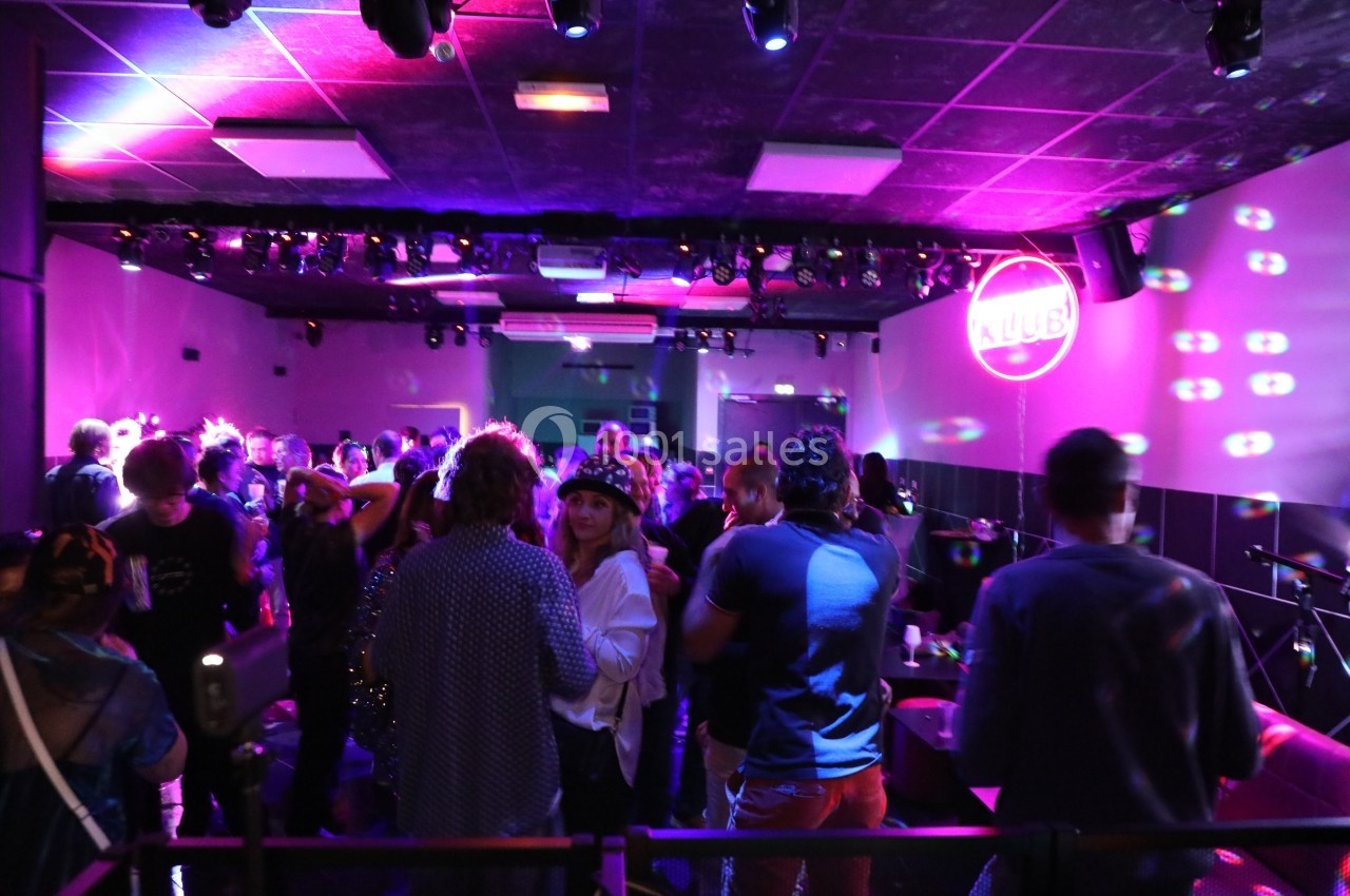 Le Club Personnes dansant et discutant dans une salle de club éclairée par des lumières colorées et un logo néon au mur.