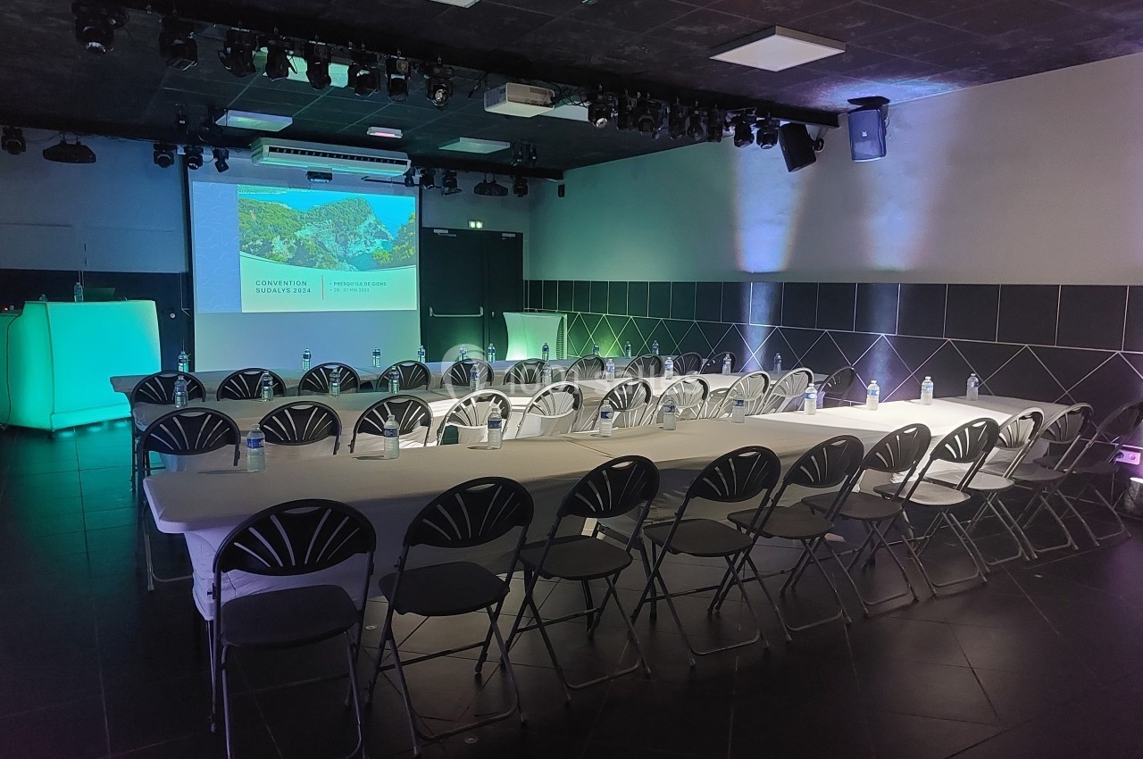 Le Club Salle de conférence sombre avec des rangées de chaises et une table, éclairée par des lumières colorées et un projecteur.