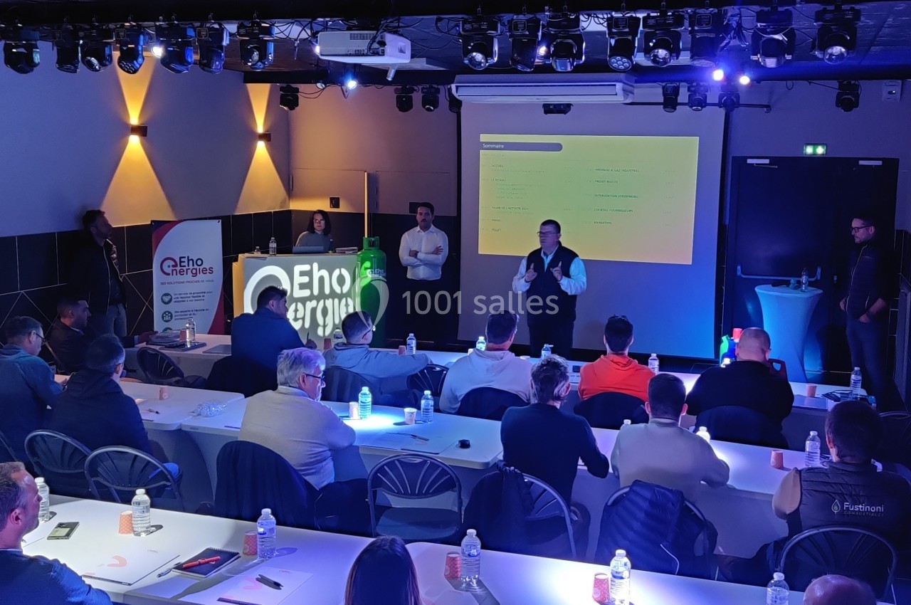 Le Club - séminaire Un conférencier s'adresse à un public assis dans une salle éclairée, avec un écran de présentation en arrière-plan.