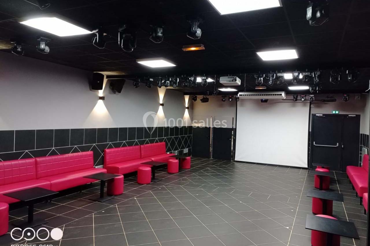 Le Club Salle moderne avec banquettes rouges, tables noires, éclairage au plafond et un écran blanc au fond.