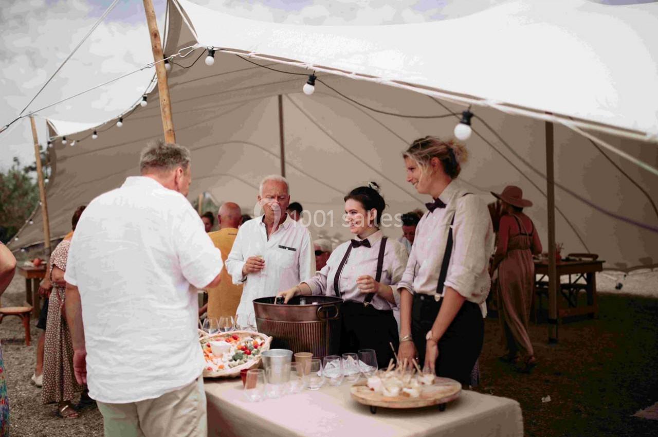 Mariage sur le terrain Les Pieds dans l'Eau Des serveurs en tenue formelle servent des boissons à des invités sous une tente blanche lors d'un événement en extérieur.