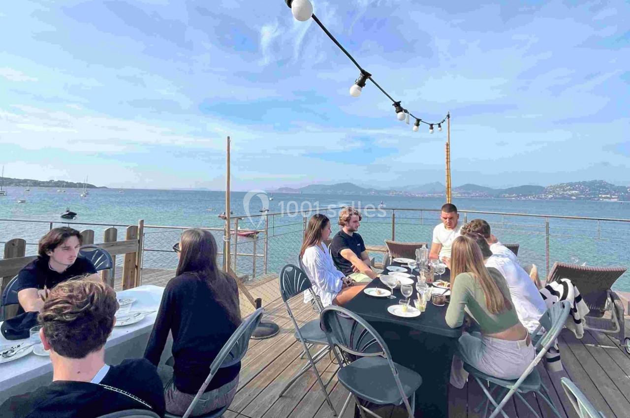 Pointe Panoramique Des personnes dînent en terrasse en bord de mer, avec une vue sur l'eau et les montagnes à l'horizon.