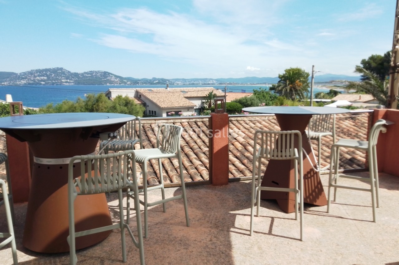 Rooftop vue mer Terrasse avec tables hautes, chaises en métal et vue sur des toits, la mer et des collines sous un ciel dégagé.