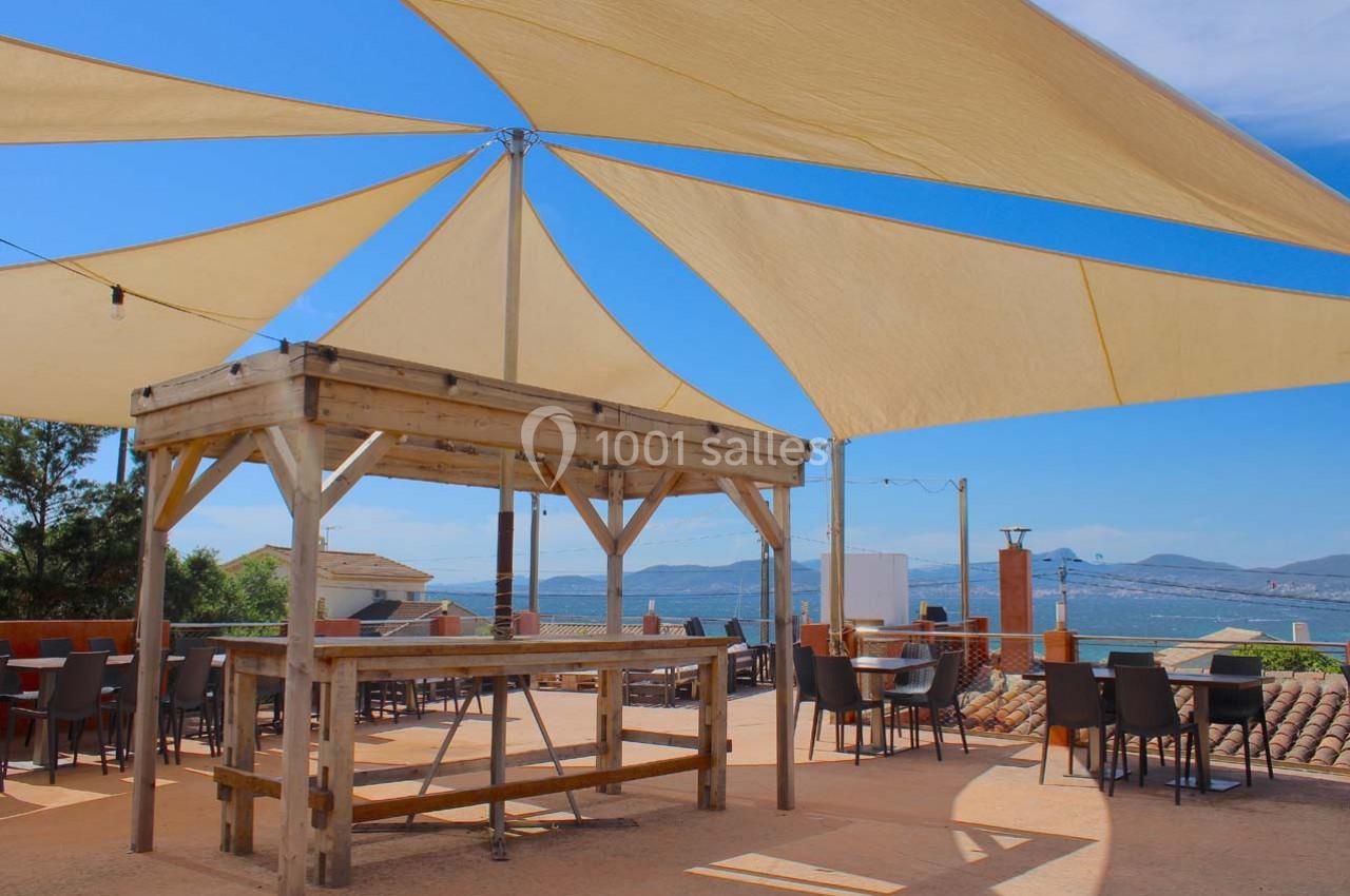 Rooftop vue mer Terrasse ensoleillée avec voiles d'ombrage, tables et chaises, offrant une vue sur la mer et les collines au loin.