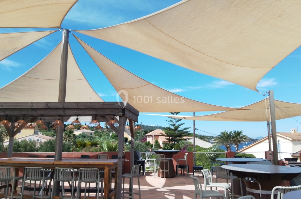 Rooftop vue mer Terrasse ensoleillée avec voiles d'ombrage, tables et chaises, entourée de végétation et maisons en arrière-plan.