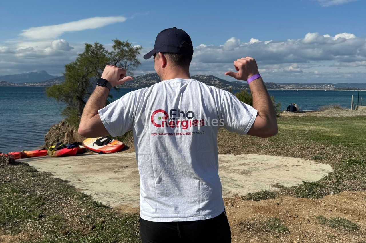 Teambuilding sur le terrain Les Pieds dans l'Eau Un homme de dos portant un t-shirt blanc avec un logo, debout sur une pelouse près d'un lac par temps ensoleillé.