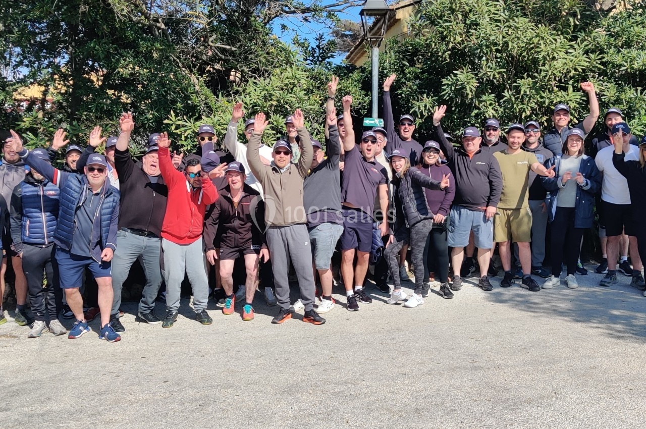 Teambuilding Un groupe de personnes souriantes levant les bras, rassemblées en extérieur devant des arbres et des buissons.