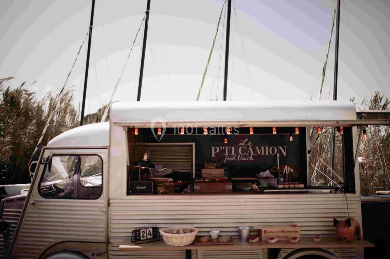 Terrain les Pieds dans l'Eau Food truck vintage stationné devant des roseaux et des mâts de bateau, avec une enseigne ’Le P'ti Camion’.