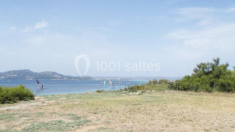 Terrain les Pieds dans l'Eau Vue d'une plage avec des planches à voile sur l'eau, des collines en arrière-plan et de la végétation au premier plan.