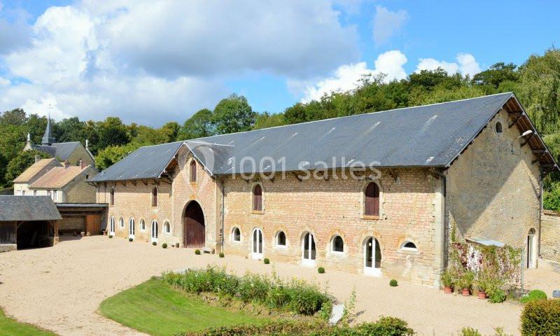 Bâtiment en pierre avec toit en ardoise, grandes arches et fenêtres, entouré d'une cour gravillonnée et de verdure.