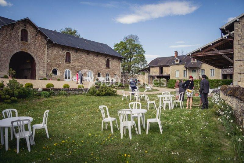 Cour extérieure d'un domaine rural avec des bâtiments en pierre, des tables blanches et quelques personnes en discussion.