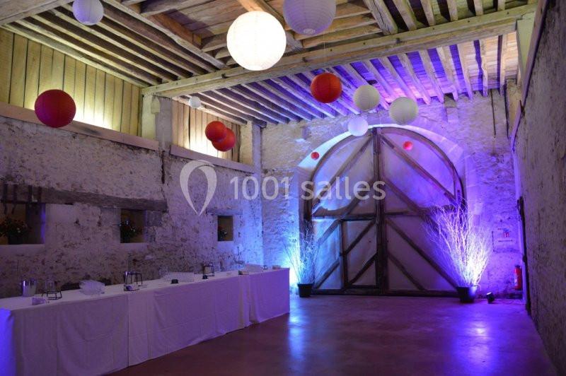 Salle en pierre avec poutres apparentes, décorée de lanternes suspendues et éclairages colorés, table dressée sur le côté.