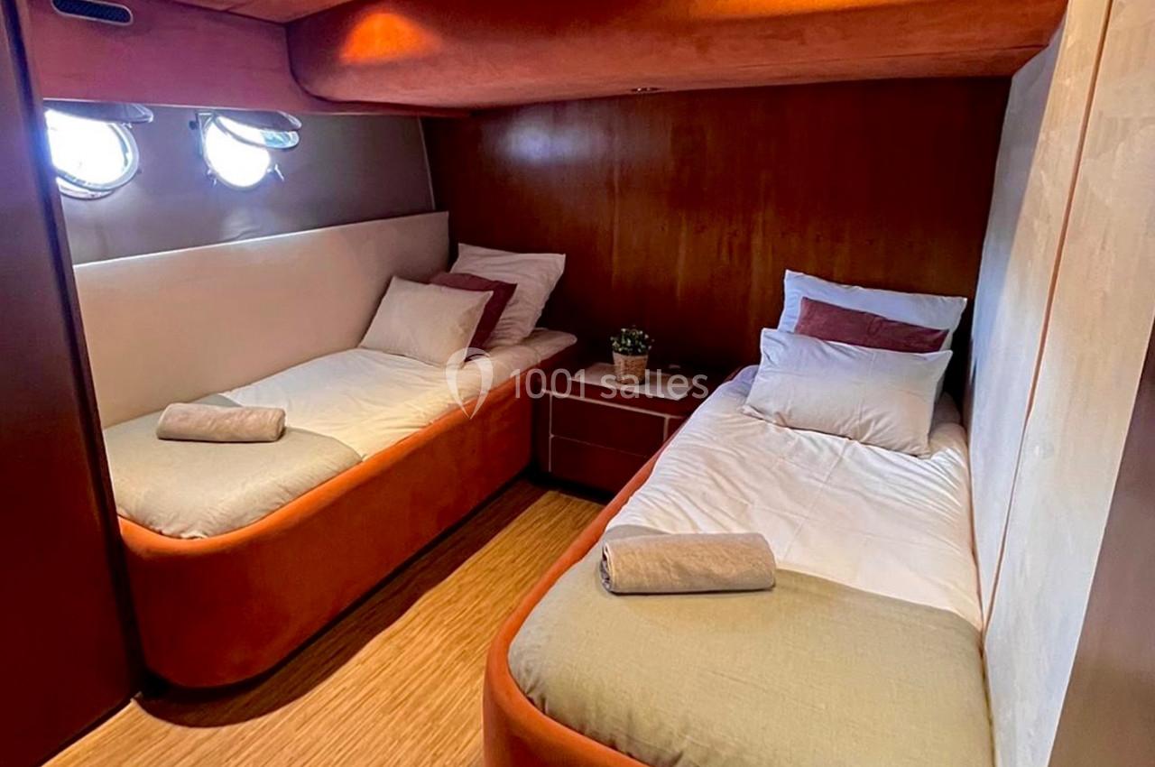 Cabine de bateau avec deux lits simples, linge de lit beige et rouge, éclairage intégré et hublots.