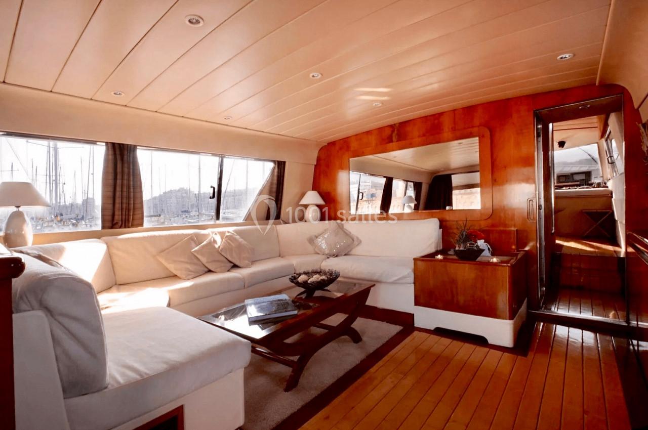 Intérieur d'un yacht avec salon spacieux, canapé blanc, table basse en bois et grandes fenêtres offrant une vue sur le port.