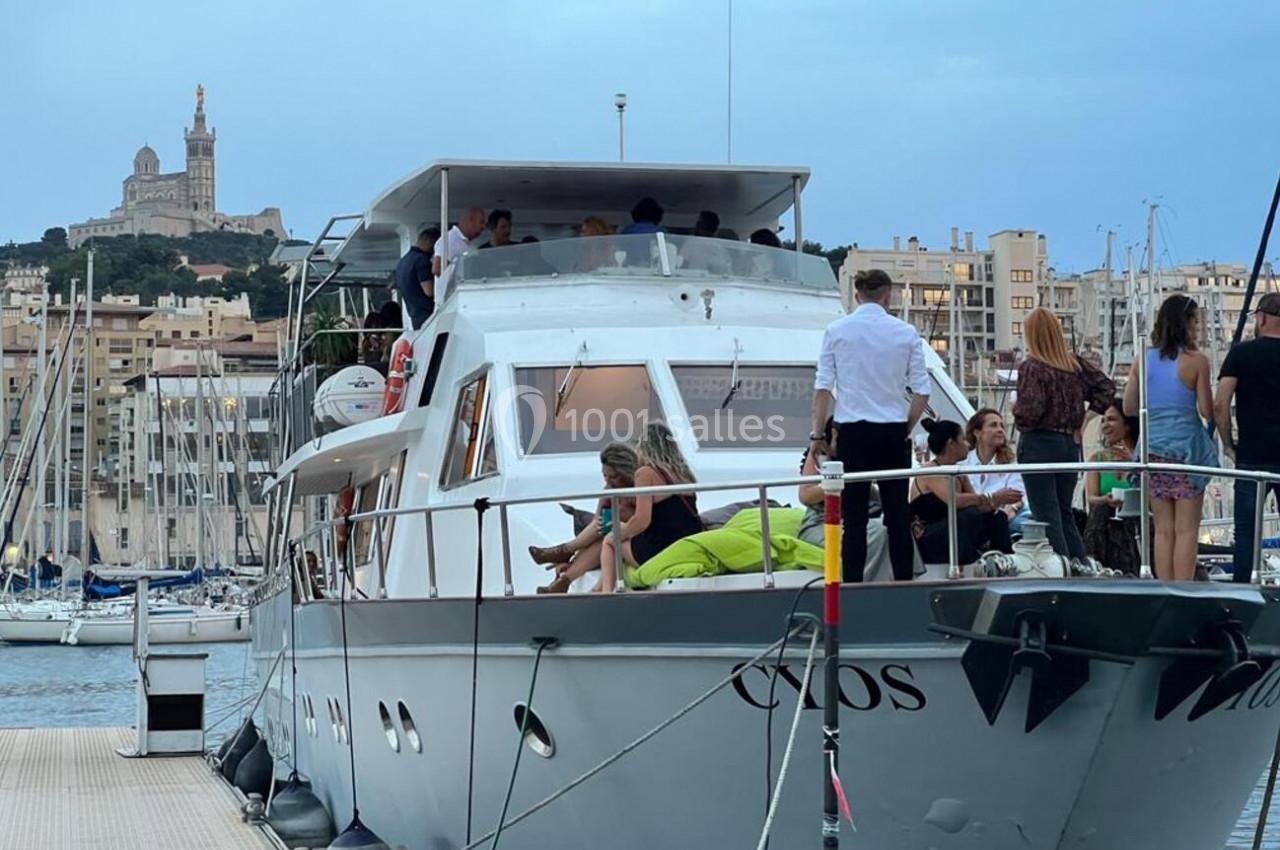 Un yacht amarré dans un port, avec des personnes à bord et en arrière-plan une basilique sur une colline.