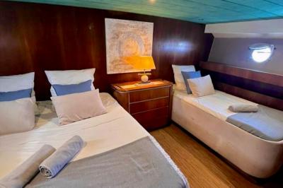 Cabine de bateau avec deux lits simples, linge de lit beige et rouge, éclairage intégré et hublots.