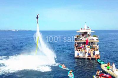 Un bateau à moteur tracte une bouée gonflable sur une mer calme, avec un yacht et une côte boisée en arrière-plan.