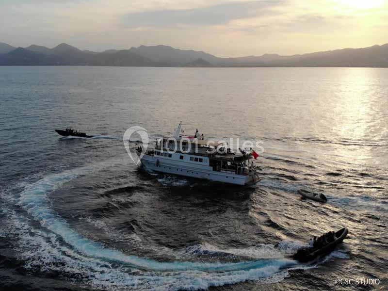 Un bateau navigue sur une mer calme entouré de petites embarcations, avec des montagnes en arrière-plan au coucher du soleil.