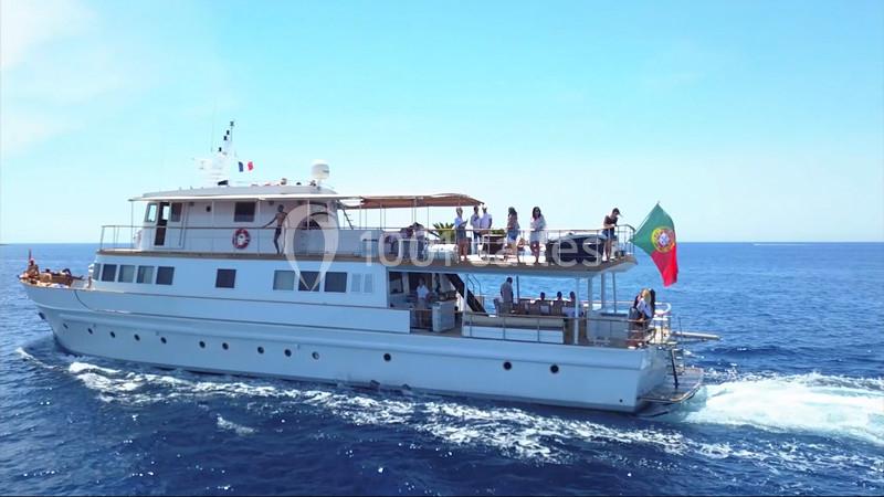 Un yacht blanc navigue sur une mer calme, avec des passagers à bord et un drapeau portugais à l'arrière.