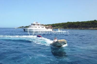 Un bateau à moteur tracte une bouée gonflable sur une mer calme, avec un yacht et une côte boisée en arrière-plan.