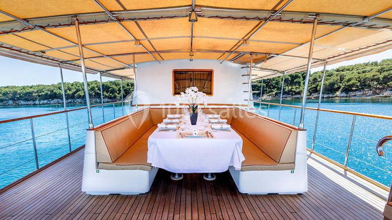 Table dressée sur le pont d'un bateau avec banquettes, sous un auvent, vue sur la mer et une forêt en arrière-plan.