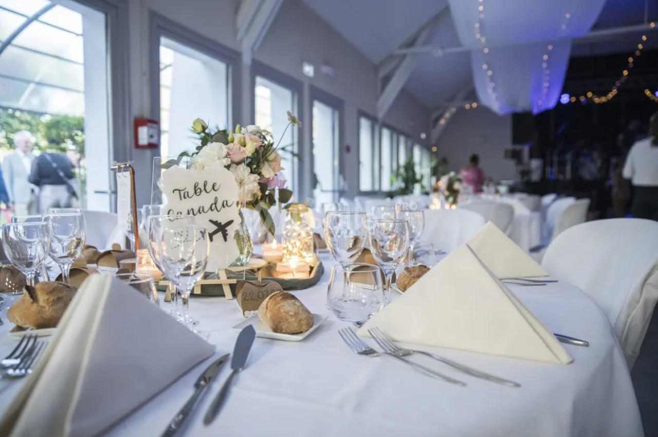 Salle de réception décorée avec des tables dressées, nappes blanches, fleurs, bougies et marque-places élégants.