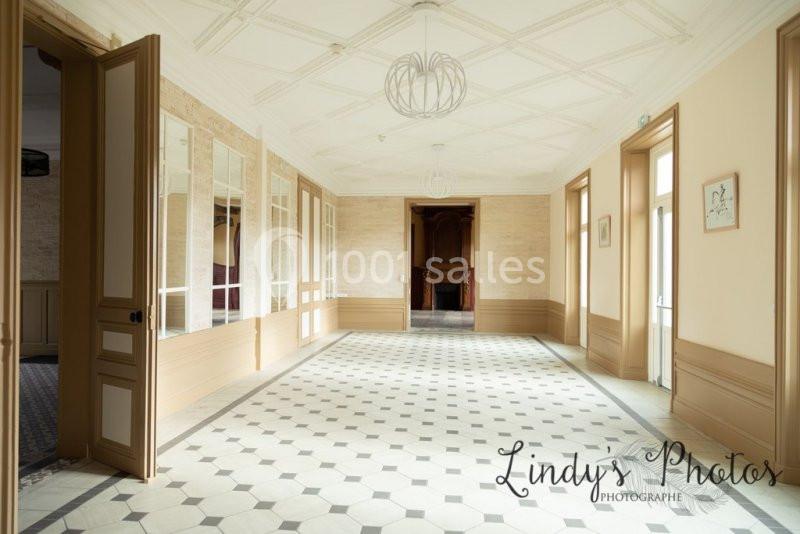 Couloir lumineux avec sol en carreaux noirs et blancs, murs clairs, portes ouvertes et cheminée en arrière-plan.