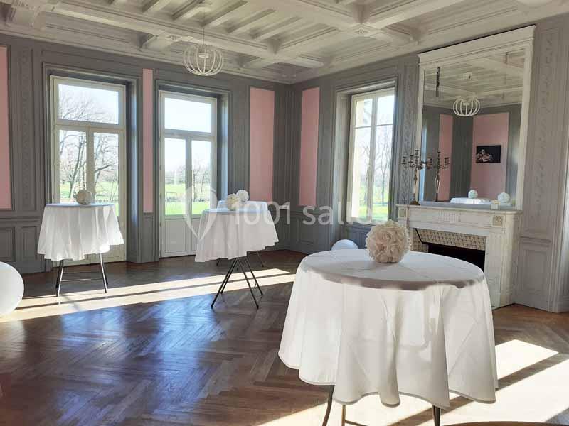 Salle lumineuse avec parquet, tables hautes nappées de blanc, grandes fenêtres et cheminée décorative.