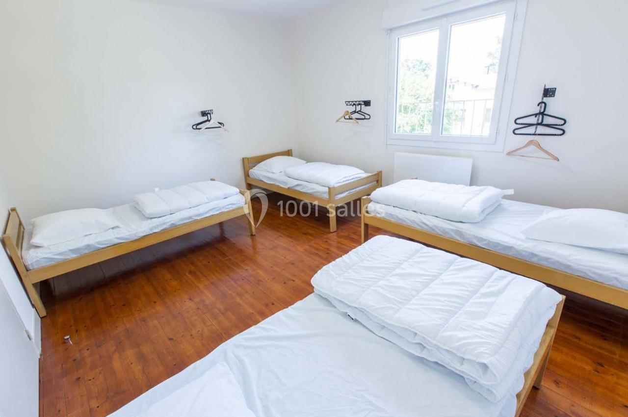 Chambre lumineuse avec quatre lits simples, draps blancs, parquet en bois et cintres accrochés au mur.