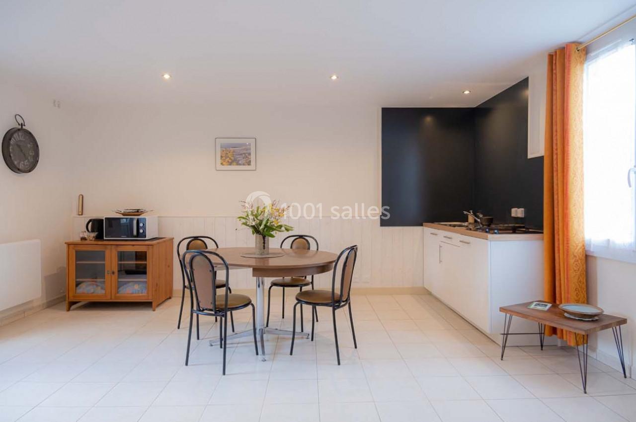 Salle à manger avec table ronde, chaises noires, cuisine équipée en arrière-plan et lumière naturelle provenant d'une…