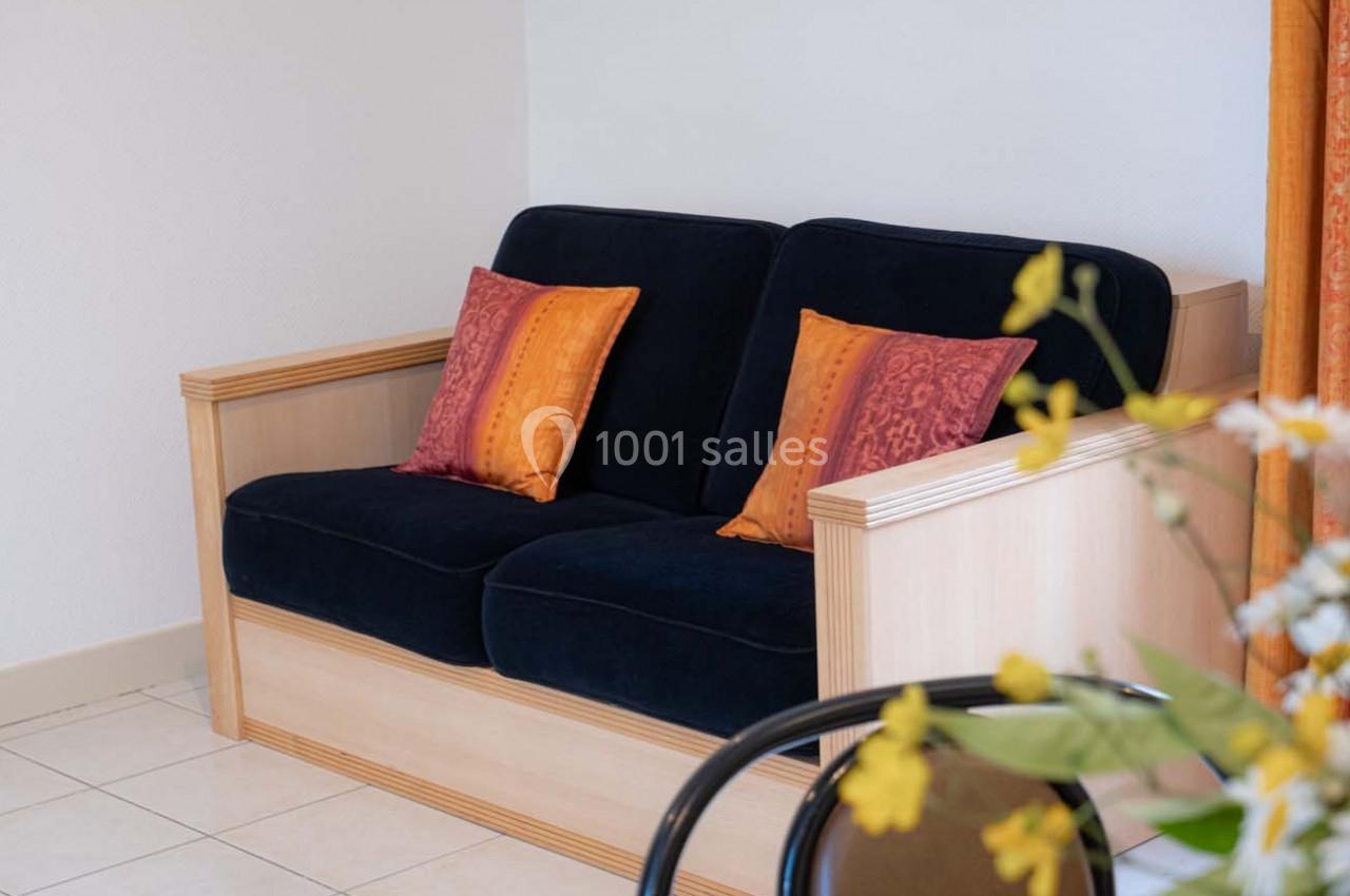 Canapé bleu à deux places avec accoudoirs en bois, décoré de coussins orange, dans une pièce lumineuse.