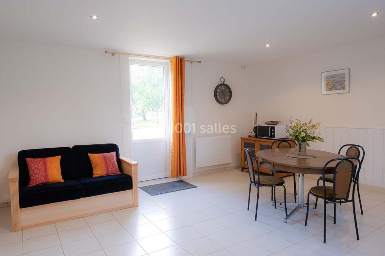 Salon lumineux avec canapé, table ronde, chaises, micro-ondes et fenêtre avec rideaux orange donnant sur l'extérieur.