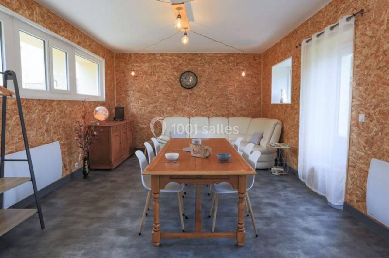Salle à manger avec table en bois, chaises blanches, canapé beige, murs en bois aggloméré et lumière naturelle.
