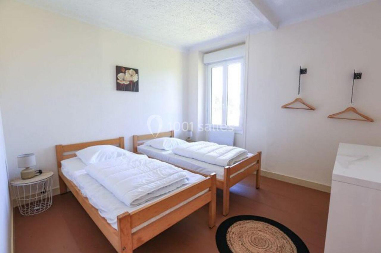 Chambre lumineuse avec deux lits simples en bois, murs blancs, fenêtre, table de chevet et cintres accrochés au mur.